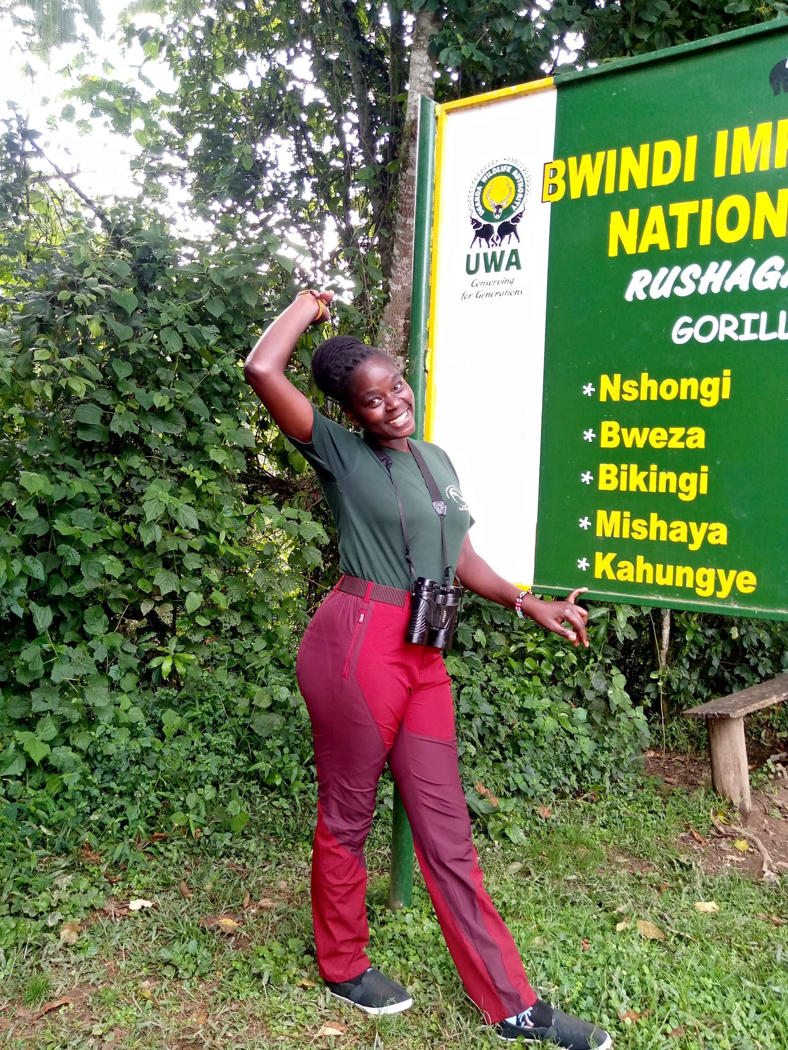 A cheerful Orugano Safaris guide stands at the entrance sign of Bwindi Impenetrable National Park, Rushaga sector