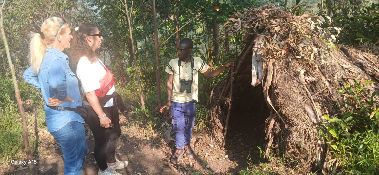 A man shows two visitors a traditional hut made of grass and leaves during a Batwa community visit